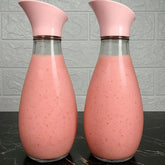 Two clear glass carafes with pink flip-top lids, filled with a pink beverage, set on a dark marble surface against a light gray textured wall.
