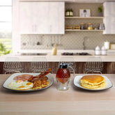 A breakfast table setup in a modern kitchen setting, featuring a plate of eggs, bacon, and hash browns on the left, a glass bottle of syrup in the center, and a plate of pancakes on the right, all arranged on a light wood table with blurred kitchen chairs and cabinets in the background.