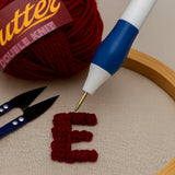 Red yarn ball with a crochet hook, scissors, and a partially completed letter 'E' on fabric.