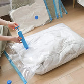 A person using a blue manual pump to evacuate air from a white quilted cushion inside a clear plastic vacuum bag with blue trim, set on a light wood floor. More vacuum bags are stacked in the background.