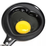 A heart-shaped black non-stick pan with a raw egg yolk and clear egg white inside, shown from a top-down view on a white background.