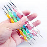 A hand holding a set of six clear acrylic nail art tools with colorful diamond-patterned handles in yellow, green, blue, pink, orange, and white, each tipped with a silver metal needle.