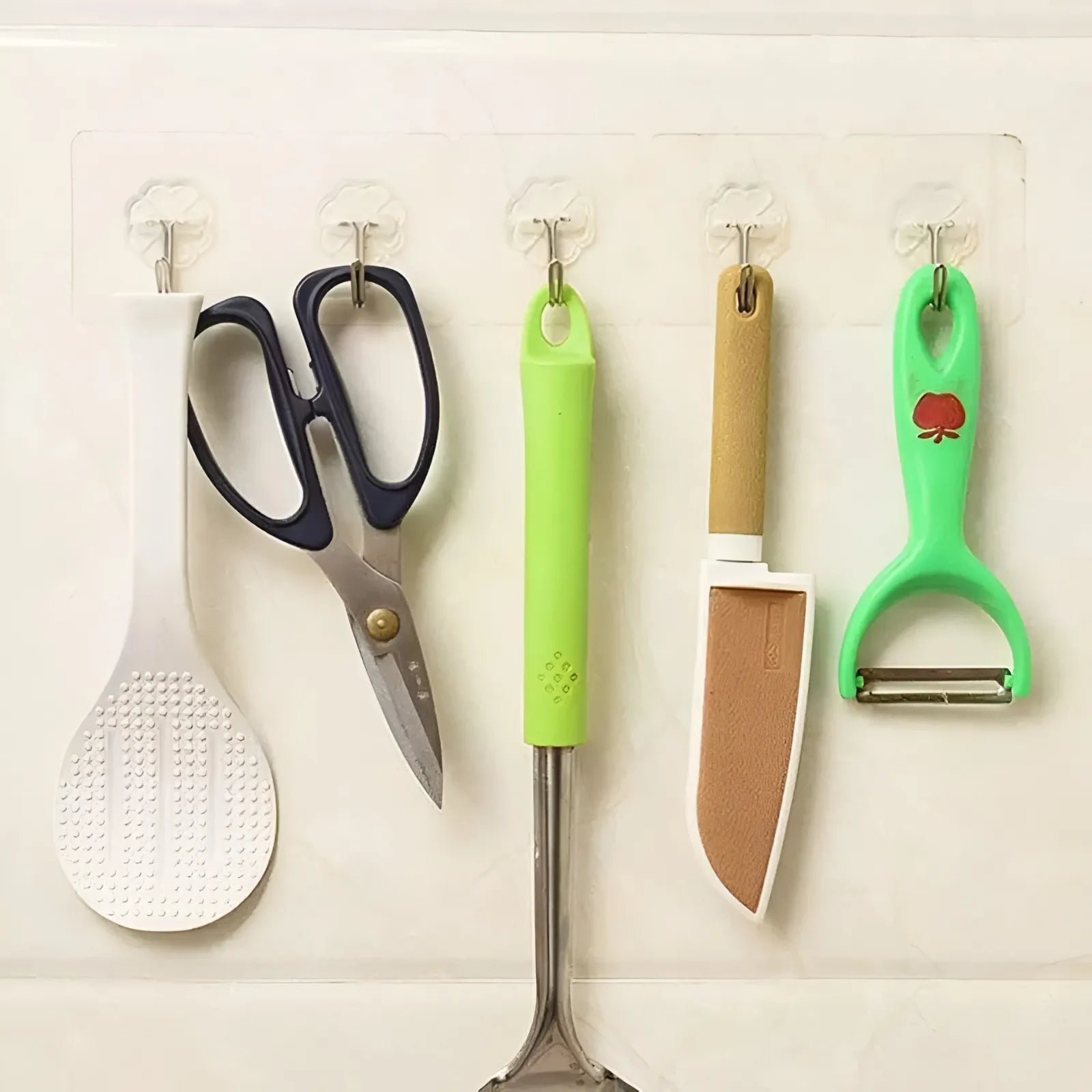 Five kitchen utensils hanging from clear wall hooks: a white ladle, dark blue scissors, a green-handled metal skewer, a beige-and-white knife, and a green peeler with a small red apple graphic.
