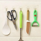 Five kitchen utensils hanging from clear wall hooks: a white ladle, dark blue scissors, a green-handled metal skewer, a beige-and-white knife, and a green peeler with a small red apple graphic.