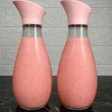 Two clear glass carafes with pink flip-top lids, filled with a pink beverage, set on a dark marble surface against a light gray textured wall.