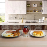 A breakfast table setup in a modern kitchen setting, featuring a plate of eggs, bacon, and hash browns on the left, a glass bottle of syrup in the center, and a plate of pancakes on the right, all arranged on a light wood table with blurred kitchen chairs and cabinets in the background.