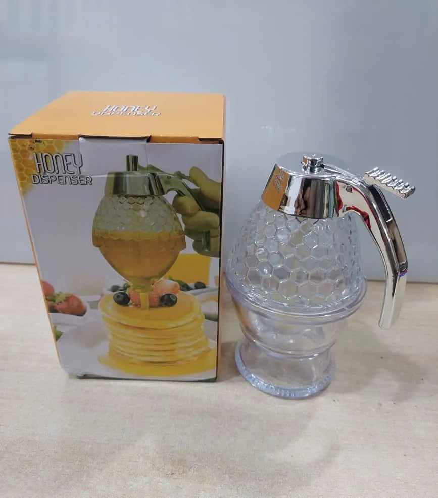 A clear honey dispenser bottle with a silver metal handle and lid, shown next to its product packaging box on a light beige surface.