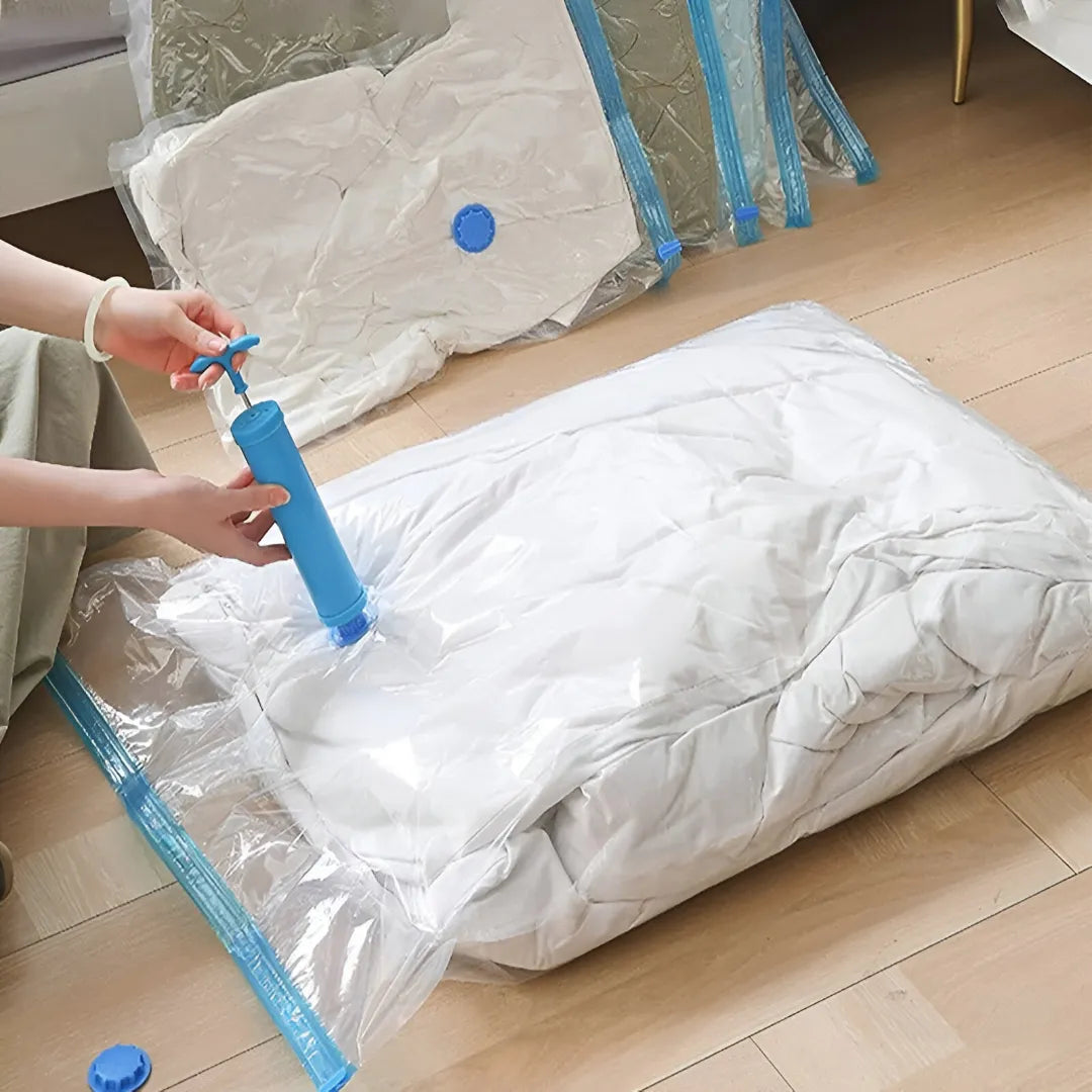 A person using a blue manual pump to evacuate air from a white quilted cushion inside a clear plastic vacuum bag with blue trim, set on a light wood floor. More vacuum bags are stacked in the background.
