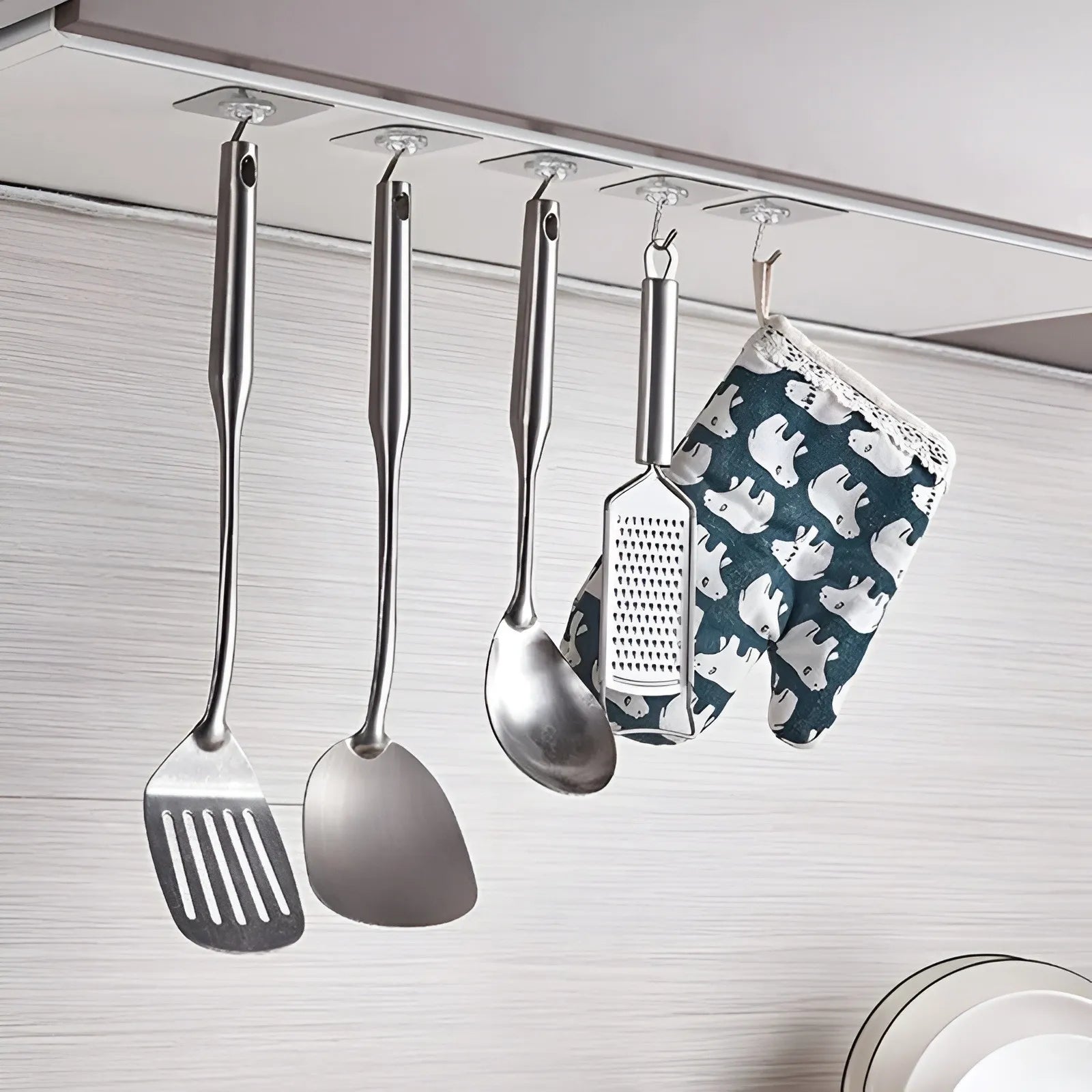 A set of stainless steel kitchen utensils, including a spatula, spoon, ladle, and grater, hanging from a metal rod on a wall, alongside a blue and white patterned oven mitt.