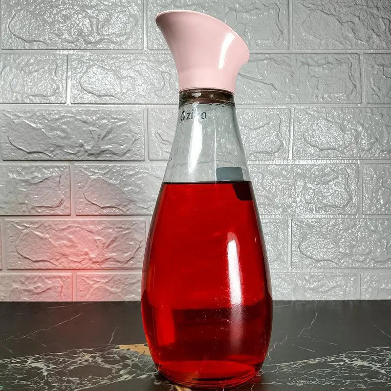 Glass carafe with a pink spout filled with red liquid on a textured surface.