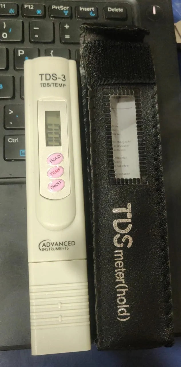 A TDS-3 TDS/TEMP meter in white with pink buttons, shown next to its black fabric carrying case on a dark keyboard background.
