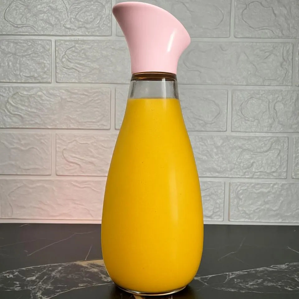 A teardrop-shaped glass carafe filled with bright yellow-orange juice, featuring a curved light pink spout and a clear glass neck, set against a white textured brick wall background.