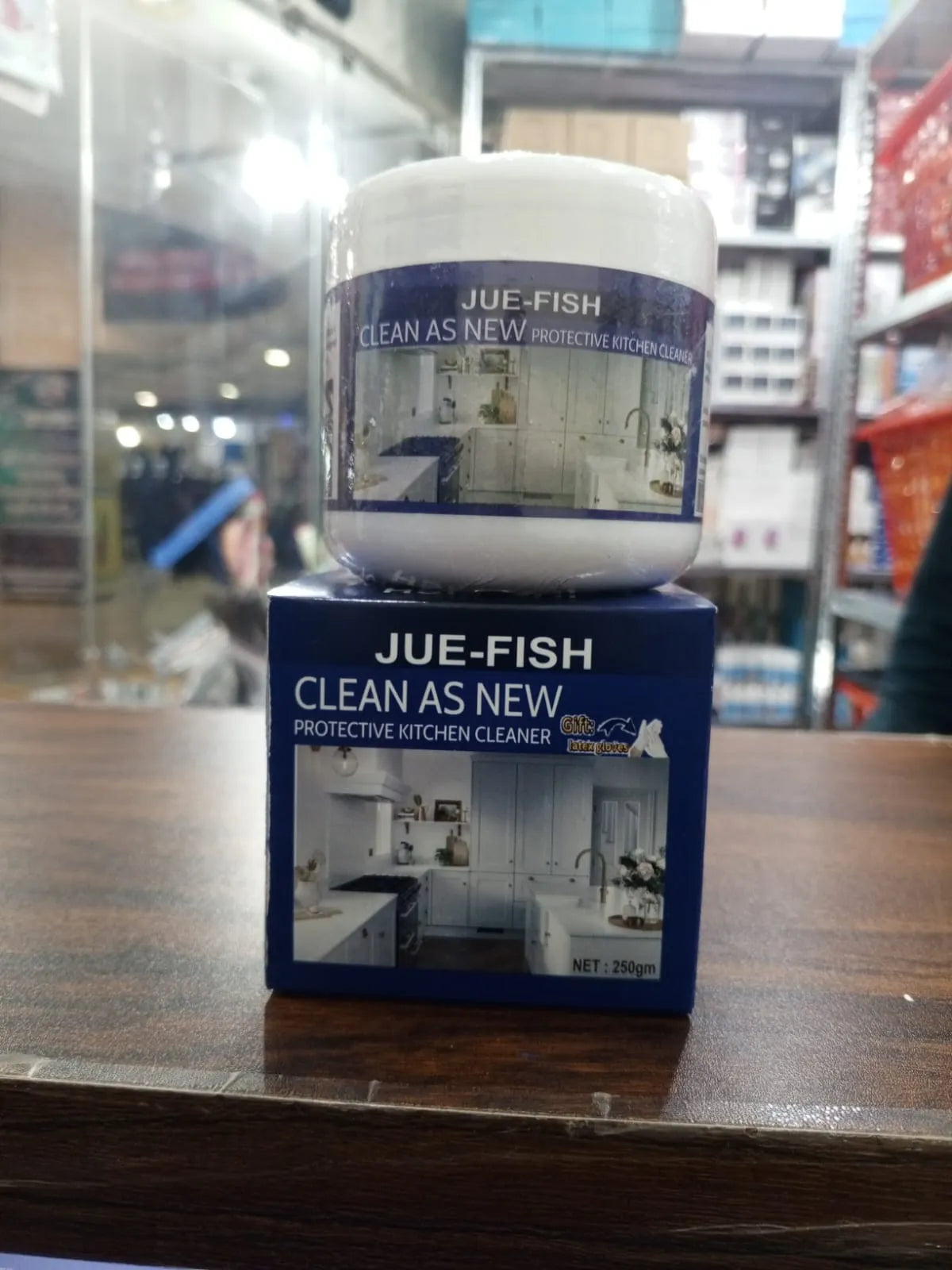 A jar of JUE-FISH Clean As New protective kitchen cleaner sits on top of its blue retail box, placed on a wooden counter in a store setting.