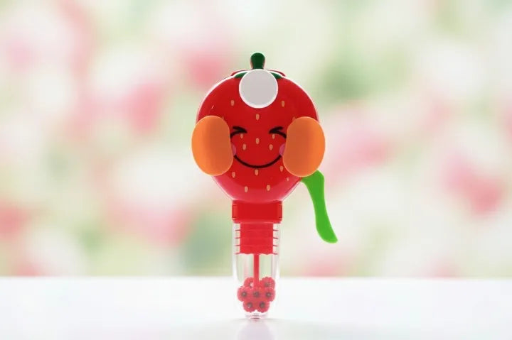 A strawberry-shaped novelty bubble pen or bubble wand with a smiling face, orange ear-like accents, a green stem, and a clear lower tube containing red bubble beads, set against a soft blurred pink and green background.
