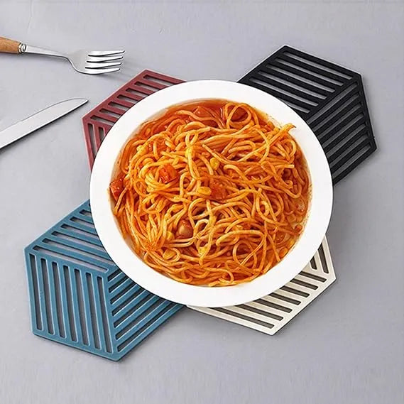 Four hexagonal silicone trivet coasters in blue, white, black, and burgundy, shown with a bowl of spaghetti on a gray tabletop.