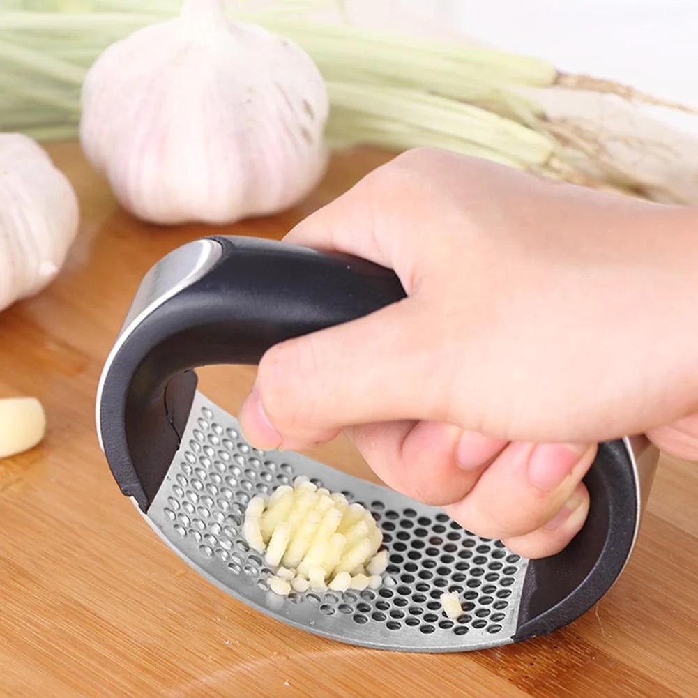 Hand using a garlic press on a wooden surface with garlic and green onions in the background.