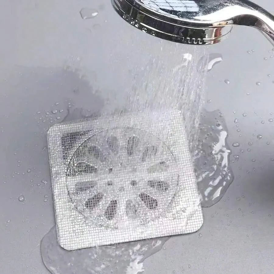 A square metal drain cover with a circular mesh pattern, shown installed in a sink under running water from a chrome shower head.
