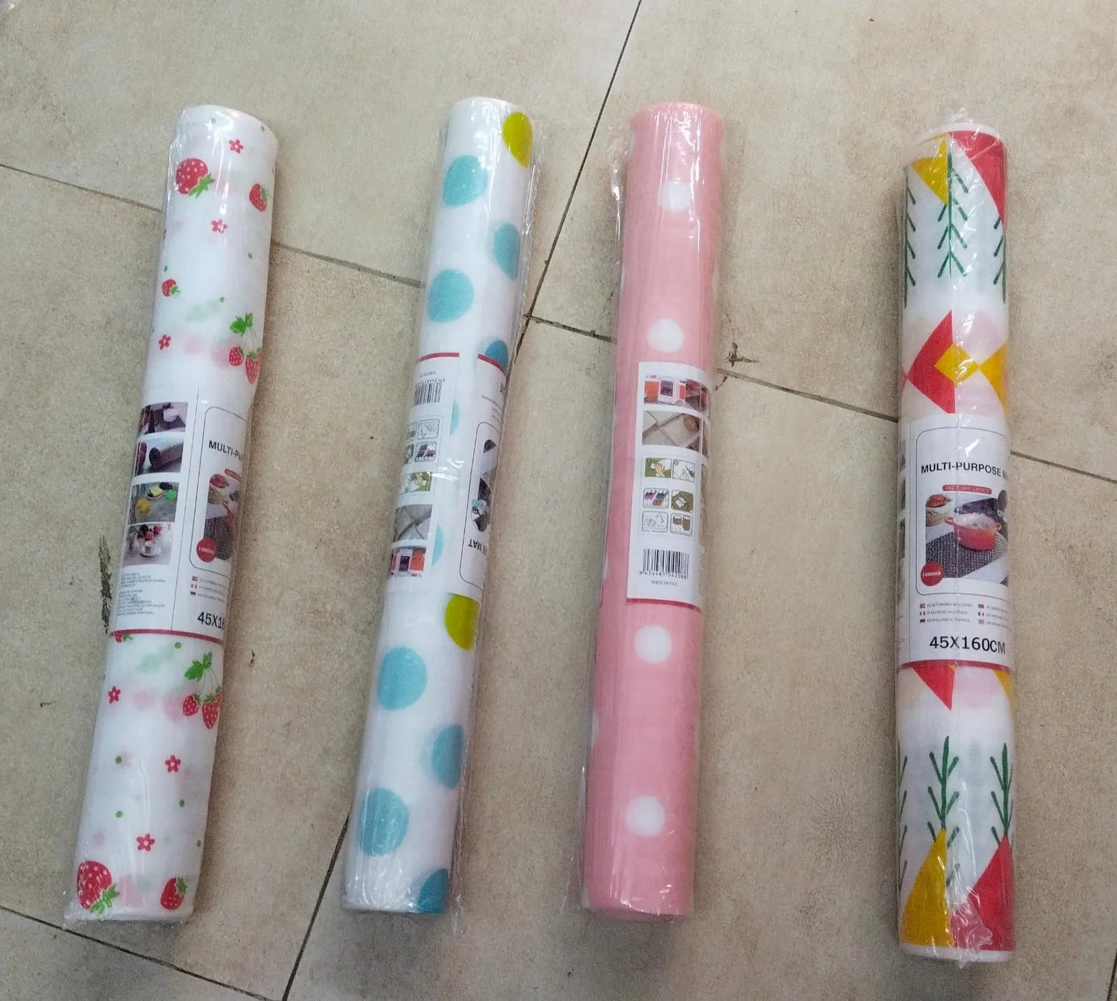 Four rolls of kitchen wrapping paper laid out on a beige tiled floor. The rolls feature different patterns: strawberry and daisy prints, blue polka dots, white polka dots, and a red, yellow, and green geometric pattern.