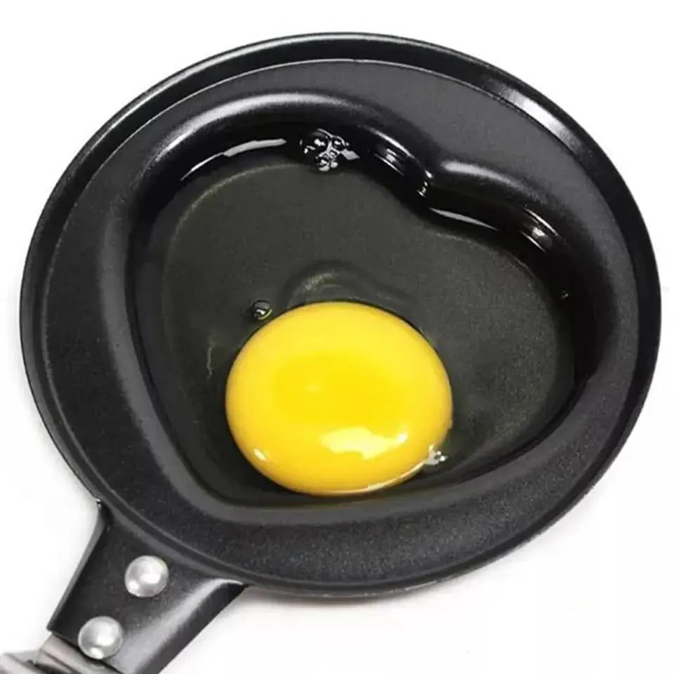 A heart-shaped black non-stick pan with a raw egg yolk and clear egg white inside, shown from a top-down view on a white background.