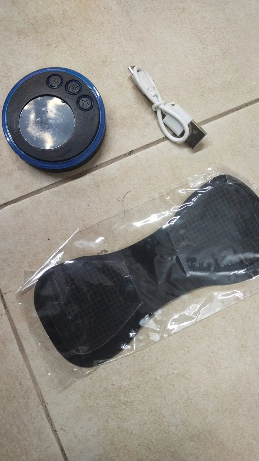 Black and blue electronic device with buttons, USB cable, and clear plastic bag on a tiled floor.