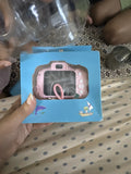 A pink toy camera in its blue retail box, held by a hand over a patterned beige and gray blanket. The box features a clear window showing the pink toy camera inside, with cartoon graphics of a dolphin and a pirate on the bottom left.