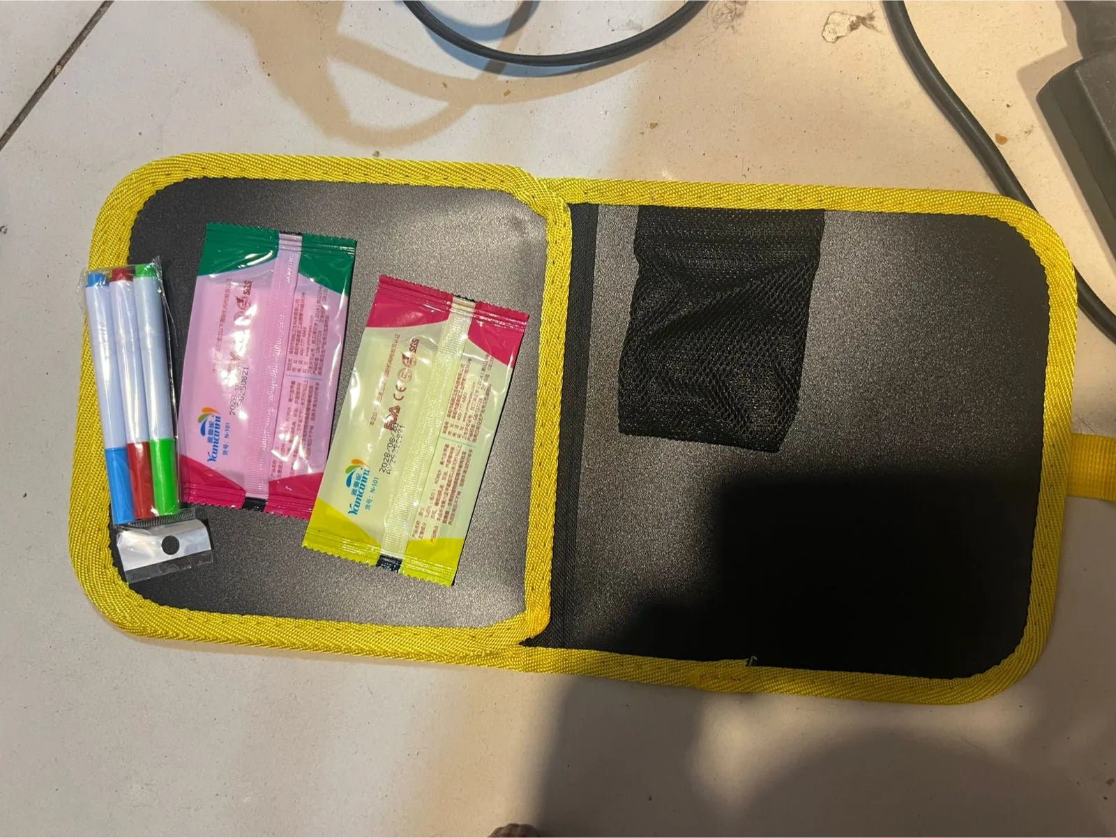 An open yellow and black emergency preparedness kit pouch containing medical supplies, including pink and green packets, a small metal tool, and a black mesh pocket, photographed on a light floor.