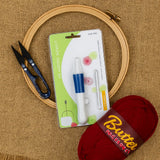 Embroidery hoop, scissors, packaging of embroidery tools, and a ball of red yarn on a textured surface.