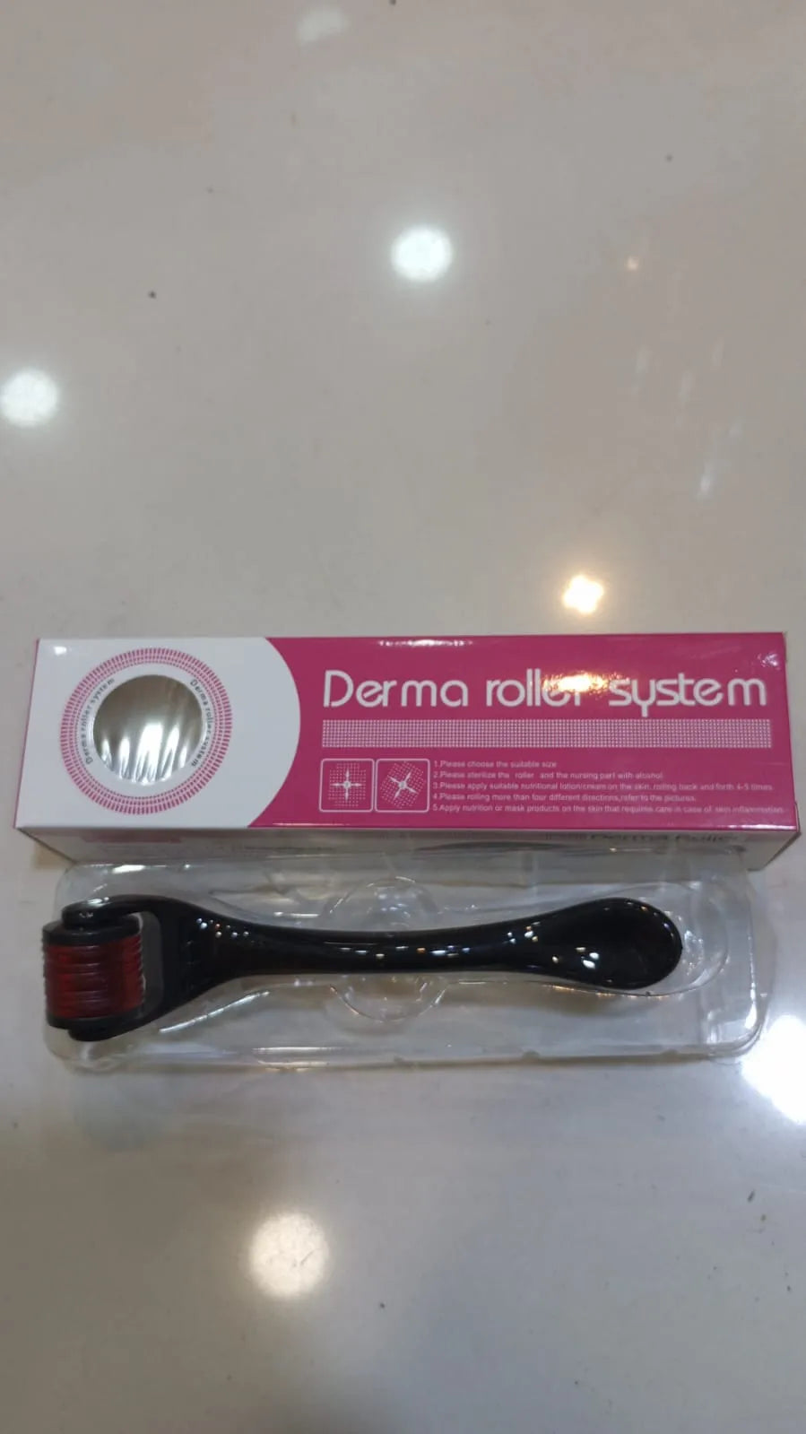 A derma roller tool in its clear plastic blister packaging with a pink and white box behind it, placed on a light-colored surface.