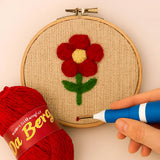 Embroidery hoop with a red flower design, red yarn, and a hand holding a needle on a beige background.