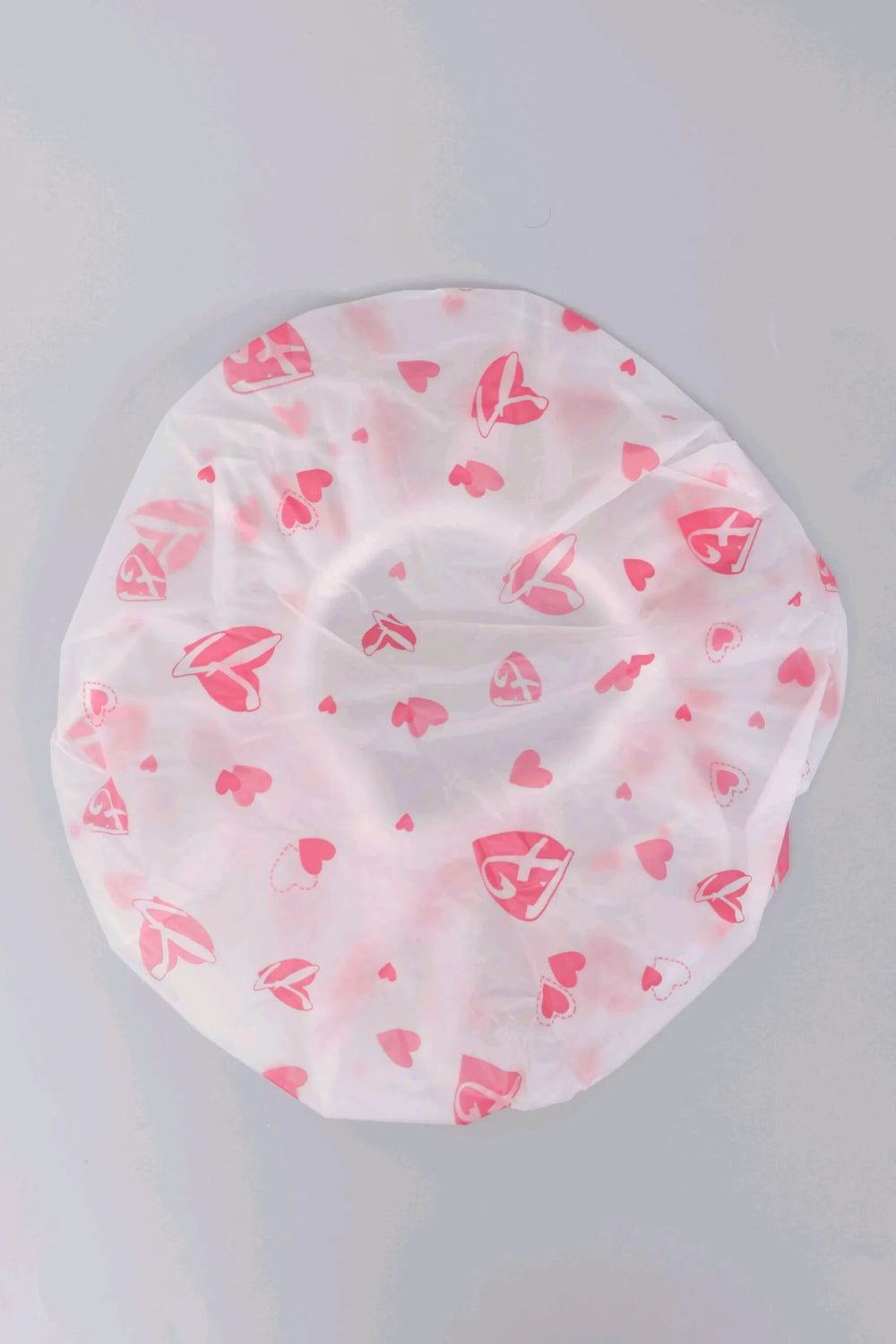 A white circular shower cap with a pink lip and heart pattern, photographed flat against a light gray background.