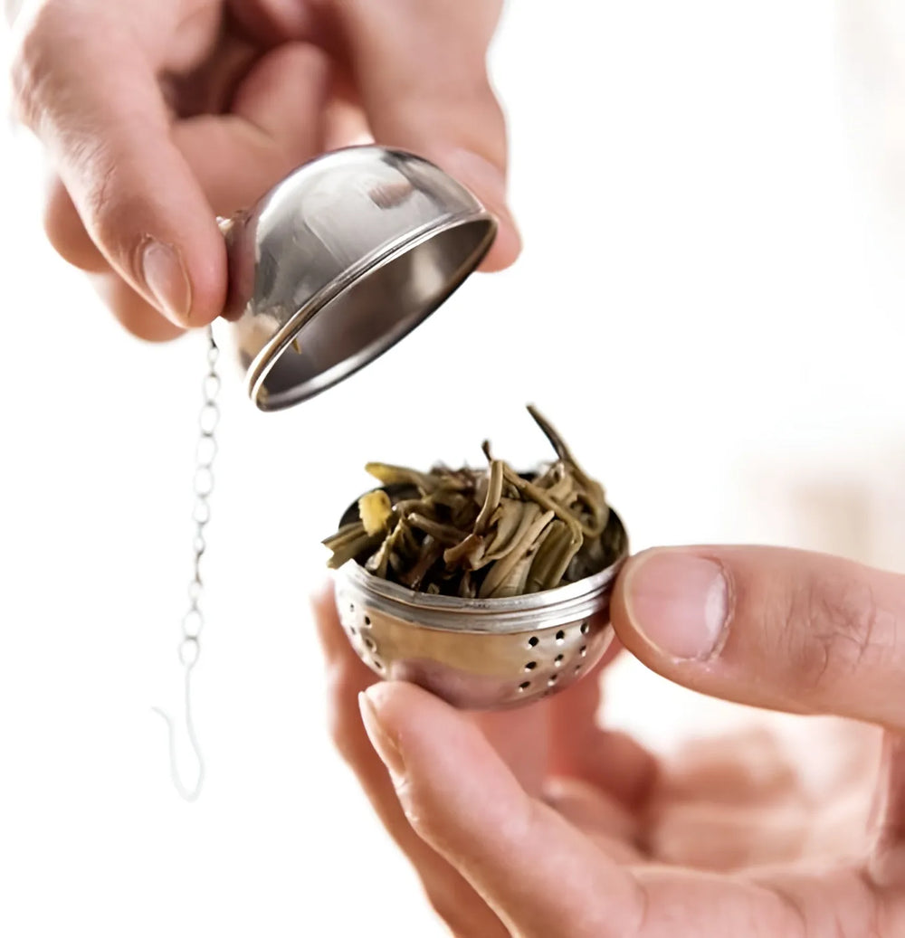 A stainless steel tea infuser ball with a hanging chain, shown being opened with one hand while containing loose tea leaves, held by another hand against a bright white background.
