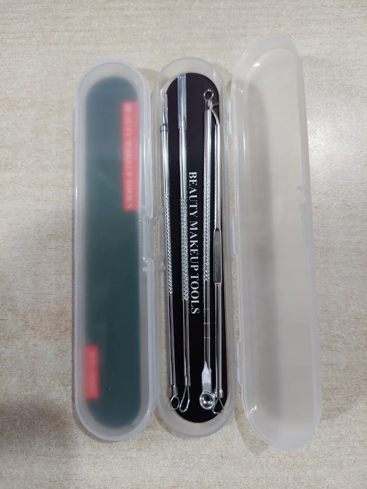 A three-piece makeup removal tool set in a clear plastic case, featuring a black nail file on the left, three silver metal picks in the center, and a translucent plastic cover on the right, shown on a light beige textured surface.