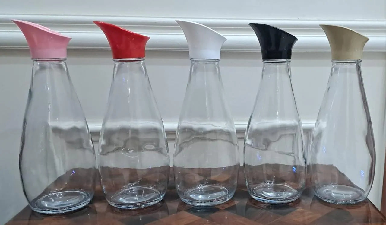 Five clear glass carafes with different colored pour spouts lined up on a wooden table against a white wall.
