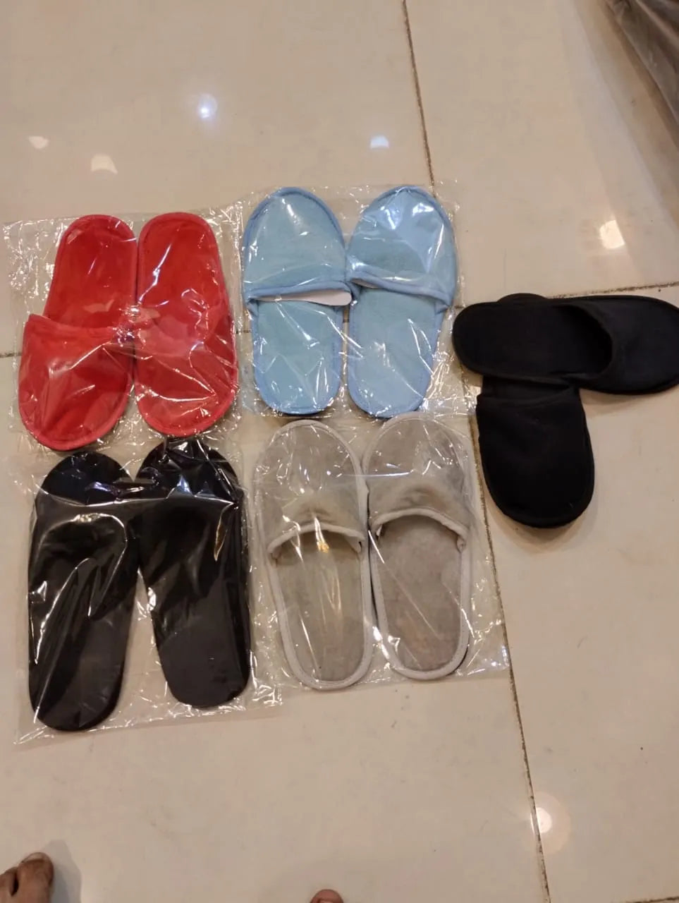 Six pairs of slip-on slippers in red, light blue, black, and gray, some still in their clear plastic packaging, arranged on a beige tiled floor.