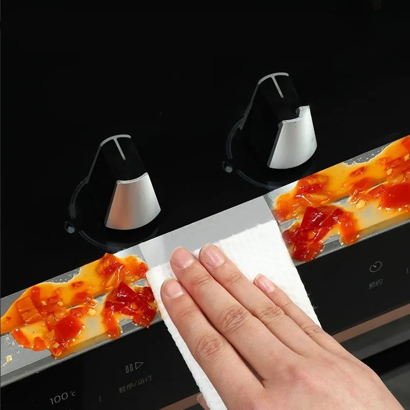 A close-up of a hand wiping a black glass cooktop with a white paper towel, with red sauce residue visible on the surface and two black-and-silver stove knobs in the background.